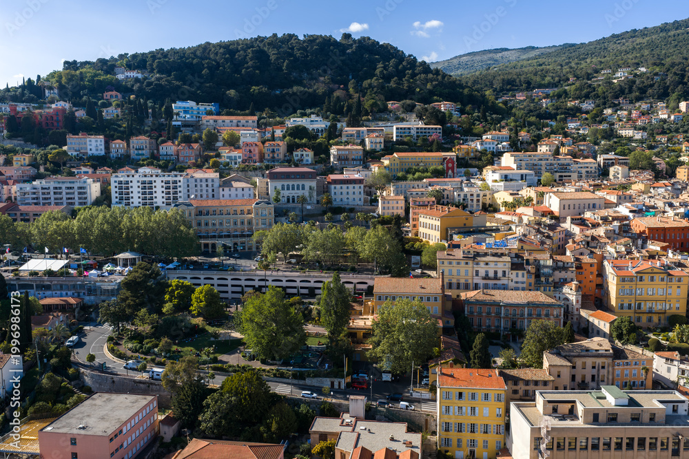 Obraz premium Aerial view of Grasse, France shows pastel buildings on slopes, terraced neighborhoods, narrow streets, and gardens under midday sun, with forested hills and a valley.