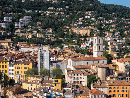 Aerial view of Grasse, France, shows terracotta rooftops on a hillside, the pale stone Grasse Cathedral tower, modern blocks above, and a scaffolded tower at midday.