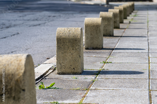 a row of weathered concrete bollards lining a paved sidewalk, featuring a perspective view that leads down a city street with long morning shadows