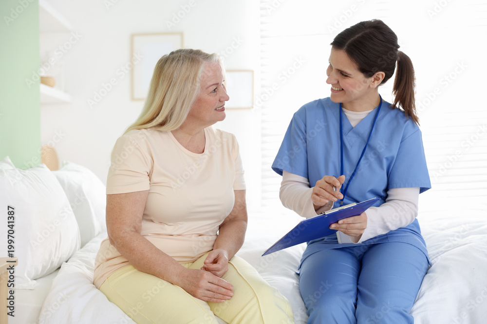Obraz premium Smiling care worker with clipboard working with senior woman on bed at nursing home