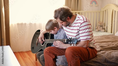Handsome dad teaches his cute son to play the guitar sitting on sofa in the living room at home. Happy Father's Day. Father teaching his child to play guitar. Kid Learning music skill. Art education