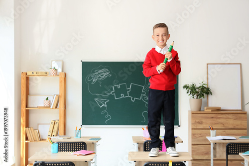 Cute little schoolboy playing with large pencil on desk in classroom