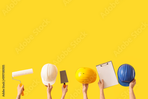 Fotografie Male hands with painter's tools, hardhats and clipboard on yellow background