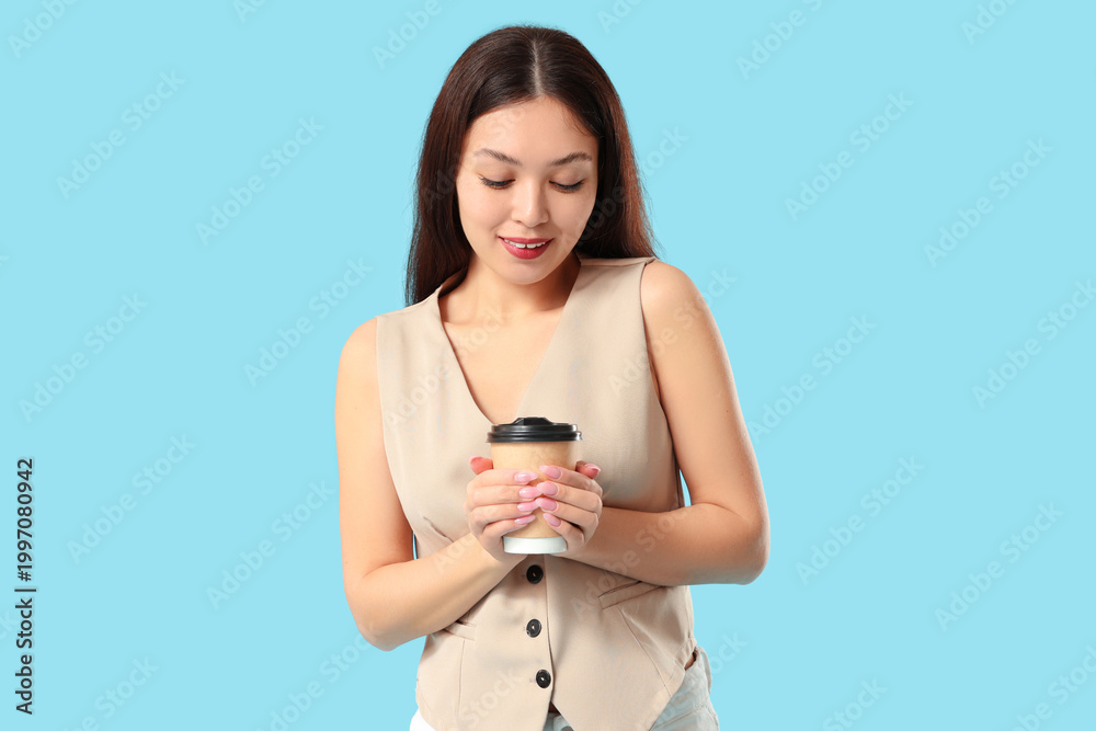 Fototapeta premium Beautiful young Asian woman in stylish beige vest with paper cup of coffee on blue background