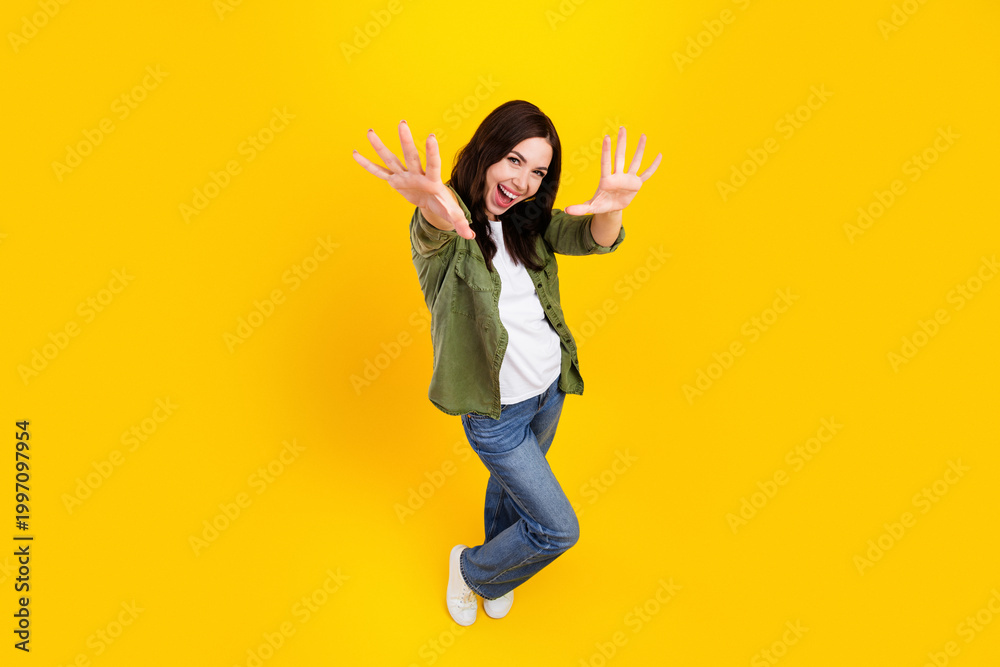 Fototapeta premium Happy young woman reaching out and laughing on a bright yellow background in casual jeans and green shirt, energetic playful pose