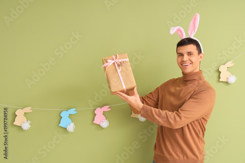 Happy young man in bunny ears with gift box and rabbits garland on green back...