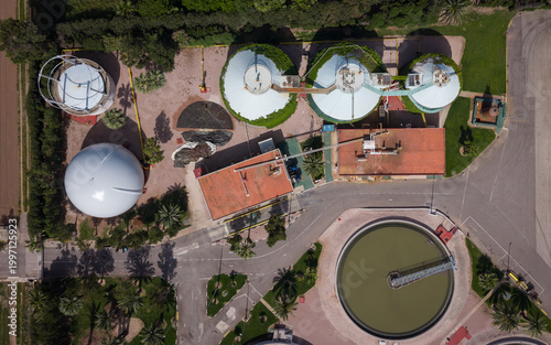 Top-down view of wastewater treatment facility showing anaerobic digestion tanks, biogas storage dome, circular clarifier, and sludge management infrastructure surrounded by palm trees