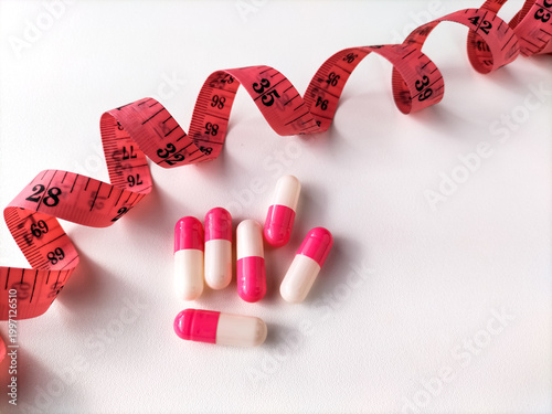 pink and white capsules are scattered on a white surface alongside a rolled-up red measuring tape. The scene suggests themes related to dieting or weight management.
