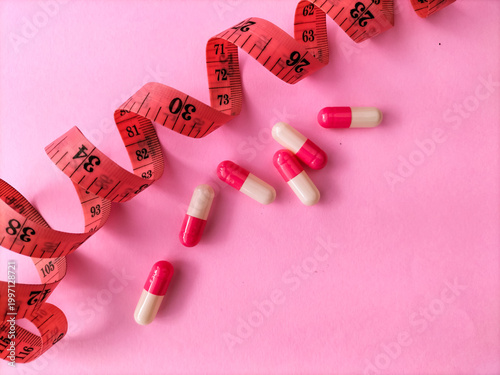 pink and white capsules are scattered on a pink surface alongside a rolled-up red measuring tape. The scene suggests themes related to dieting or weight management.