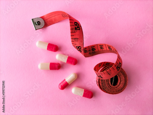 A pink background displays a red measuring tape, partly unrolled, alongside several capsules with pink and white halves. Emphasizing a weight or health-related theme.