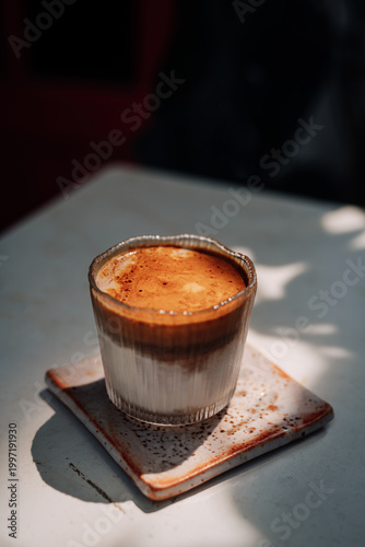 Dirty coffee in a clear glass with layered milk and espresso crema, placed on a ceramic coaster under natural light, showcasing minimalist cafe style and modern beverage aesthetics.