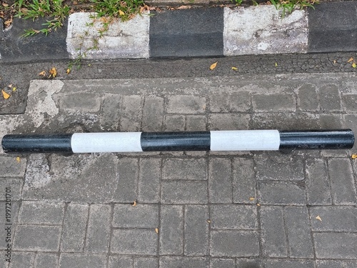 Black and white road barrier lies on brick pavement. Concrete curb with grass grows beside it. Dull autumn leaves scatter nearby. Barrier signals restricted access or parking