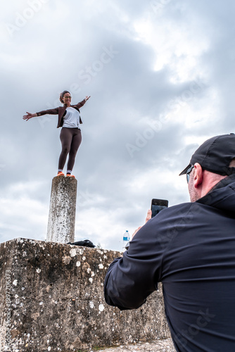 Obraz na plátne Man capturing cheerful woman standing with arms outstretched on a geodetic pilla