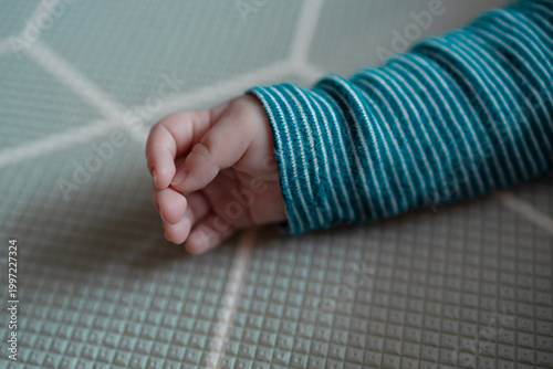 Macro shot of a tiny infant hand in a blue striped sleeve resting on a textured play mat. Detailed view of a newborn's fingers. Concept of growth, fragility, childhood, and new life.