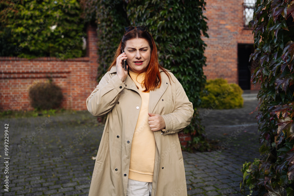 Fototapeta premium Young woman in coat talking on smartphone while standing in courtyard near brick building and greenery.
