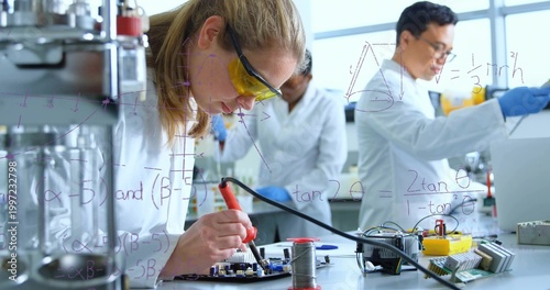 Soldering researcher in lab coat and yellow goggles working on PCB at bench, with soldering iron