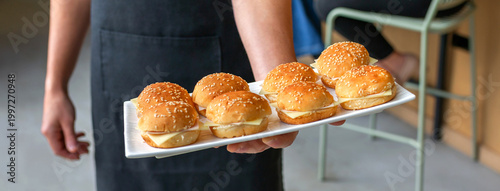 Banner of unrecognizable waiter in a black apron holding a white tray with delicious mini burgers with sesame seed buns and cheese, offering appetizer snacks at a catered event or restaurant