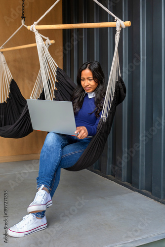 Young woman confidently working on a laptop in a comfortable hammock chair, symbolizing modern remote work flexibility and a relaxed, efficient lifestyle. Digital nomad concept.