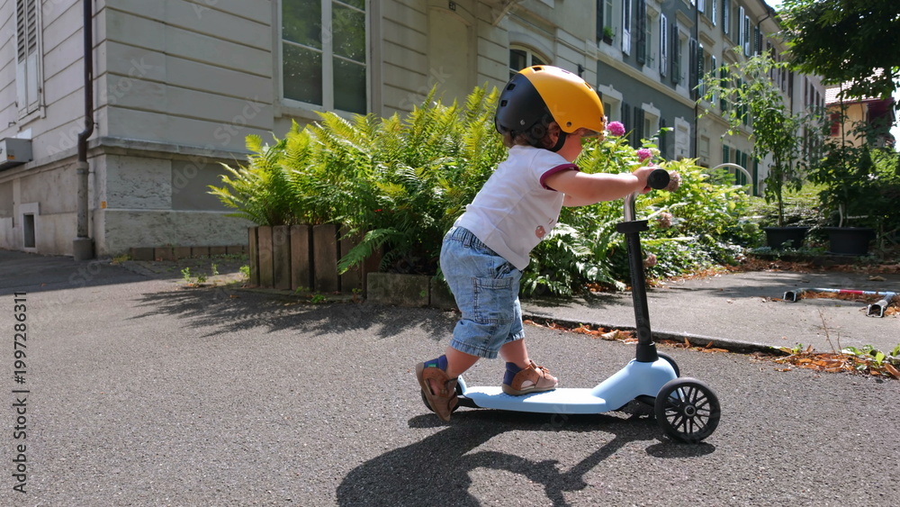 Obraz premium Toddler wearing helmet rides scooter outside residential building on sunny day, gliding along sidewalk near plants and apartment windows in suburban setting
