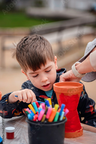 Concentrated boy painting red clay pot with brush while mother helps hold it outdoors