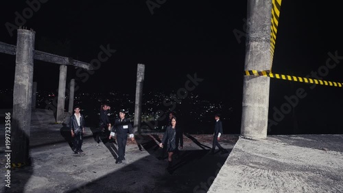 A stylishly dressed group of young people walks and poses in an unfinished concrete building overlooking the city lights at night. A cool and atmospheric shot for fashion or subculture themes.