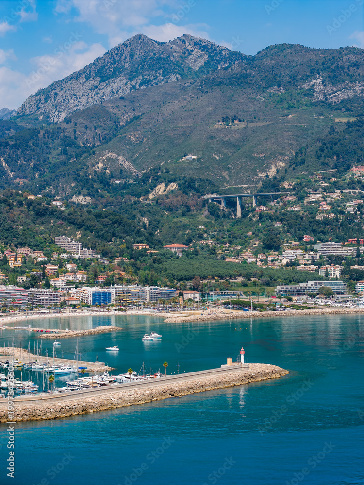 Fototapeta premium Aerial view of Menton, France shows the Old Port, marina, and turquoise bay with a stone breakwater and lighthouse, pastel town, viaduct, moored yachts, and midday light.