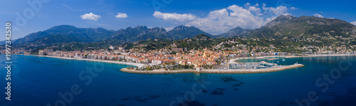 Aerial view of Menton on the French Riviera shows the Old Port, marina, promenade, and ochre bell towers by the Mediterranean and Alps, shot in clear daytime.