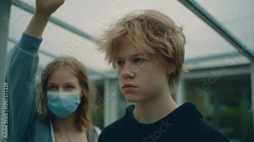 Young man with blonde hair looking forward, woman in background wearing a face mask, young adult