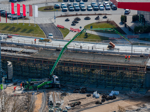 Aerial view of the Southern Bridge phase 4 in Riga, Latvia. Workers guide a long green concrete pump across a reinforced deck, with scaffolding, barriers, lots, and circles.