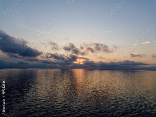 Dramatic sunset over a calm Mediterranean Sea, with golden sunlight reflecting on the water surface beneath a moody cloudscape of soft pink and blue-grey clouds, viewed from open sea.