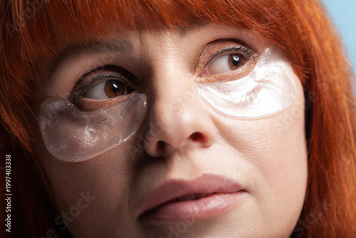 Pensive middle aged woman with vibrant red hair wearing specific anti-aging treatment with large aesthetic eye patches isolated on blue background