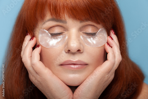 Relaxed middle aged woman with vibrant red hair and closed eyes gently resting her chin on her hands while wearing under eye patches isolated on blue background