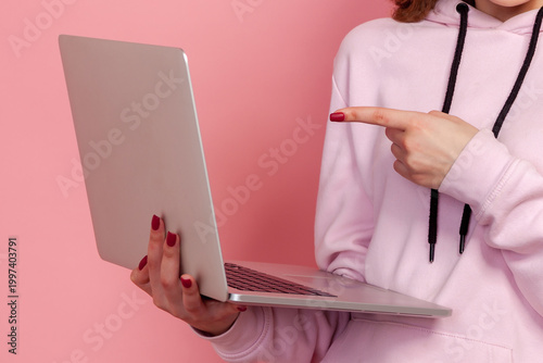 Cropped closeup view of woman holding laptop pointing at display with finger isolated on pink background