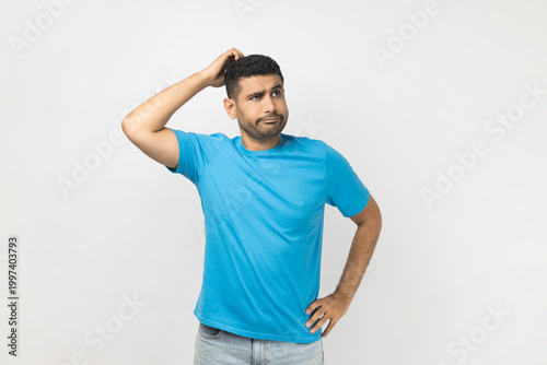 Puzzled confused unshaven man wearing blue T- shirt standing rubbing his head, looking away, thinking about something, trying to remind something. Indoor studio shot isolated on gray background.