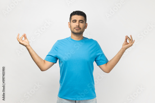 Portrait of relaxed young adult unshaven man wearing blue T- shirt standing practicing yoga, keeps eyes closed, trying to calm down. Indoor studio shot isolated on gray background.
