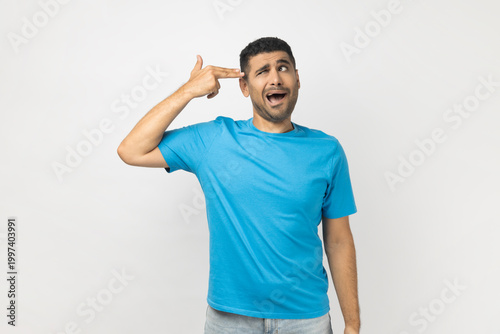 Portrait of sad depressed stressed young adult unshaven man wearing blue T- shirt standing making finger gun gesture to his head, wants to kill himself. Indoor studio shot isolated on gray background.