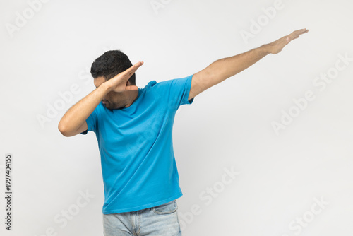 Portrait of positive excited unshaven man wearing blue T- shirt standing showing dab dance gesture, dancing, moving, internet trends. Indoor studio shot isolated on gray background.