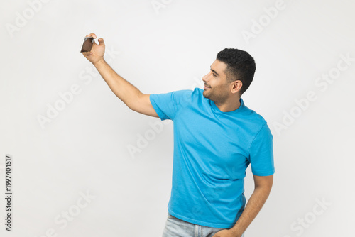 Portrait of cheerful unshaven man blogger wearing blue T- shirt standing using cell phone for broadcasting livestream, expressing positive emotions. Indoor studio shot isolated on gray background.