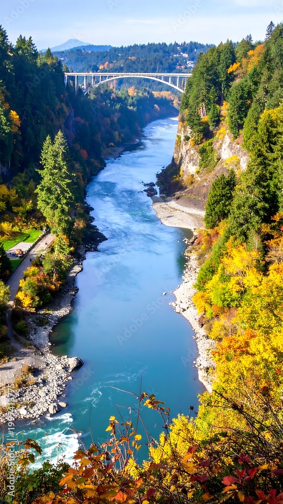 Obraz premium River flows through a gorge with autumn foliage, a bridge spans afar under a sunny sky