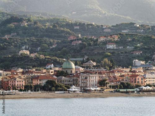 View of Albisola Superiore, Liguria, Italy, seen from the sea, with the green copper dome of the Chiesa di Nostra Signora Stella Maris dominating the waterfront, forested hillside in background
