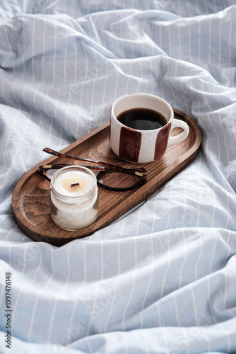 A cozy morning scene: a wooden tray on striped bedding holds a ceramic coffee cup, a lit candle in a jar, and a pair of tortoiseshell glasses the epitome of relaxed comfort.