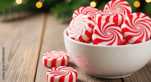 Close-up shot of red and white peppermint swirl candies in a white ceramic bowl on a rustic wooden table with festive evergreen and lights, confection, delicious, lights