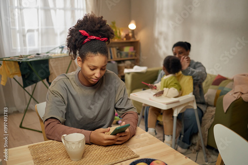 Фотография Teenage Black girl checking smartphone at table while mother caring for child in background, showing low income family life