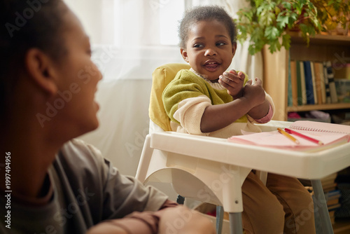 Obraz na plátně Black mother watching toddler sitting in high chair and gesturing during home learning activity, showing low income family life
