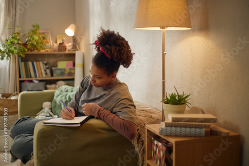 Canvas Print African American child studying at home, writing in notebook while sitting on sofa, showing low income family life and education access