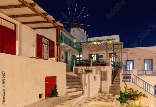 Blue hour view of traditional houses and a historic windmill in Mykonos, Greece, with whitewashed walls, colorful shutters and soft evening light in a quiet island street.
