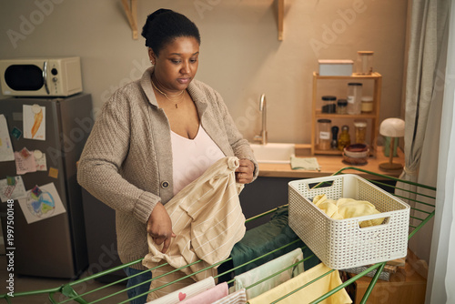 Quadro em tela Black young adult mother hanging laundry indoors, managing household chores in modest kitchen and showing financial hardship