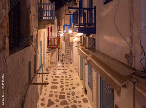 Night view of a narrow street in Mykonos, Greece, with whitewashed walls, colorful balconies, stone paving and warm lights in the quiet old town.