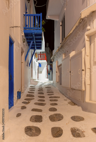 Night view of a narrow street in Mykonos, Greece, with whitewashed walls, colorful balconies, stone paving and warm lights in the quiet old town.