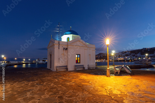 Blue hour view of a seaside church in Mykonos, Greece, with a blue dome, harbor lights, stone pavement and calm water along the waterfront.